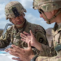 Two people wearing fatigues and helmets with camouflage have an outdoor discussion standing over a surface with documents.