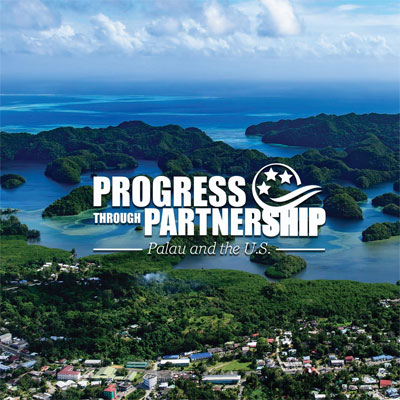 A logo reads “Progress Through Partnership Palau and the United States,” centered in a photo of a green shore with small structures on the lush ground and small islands in a large body of water.