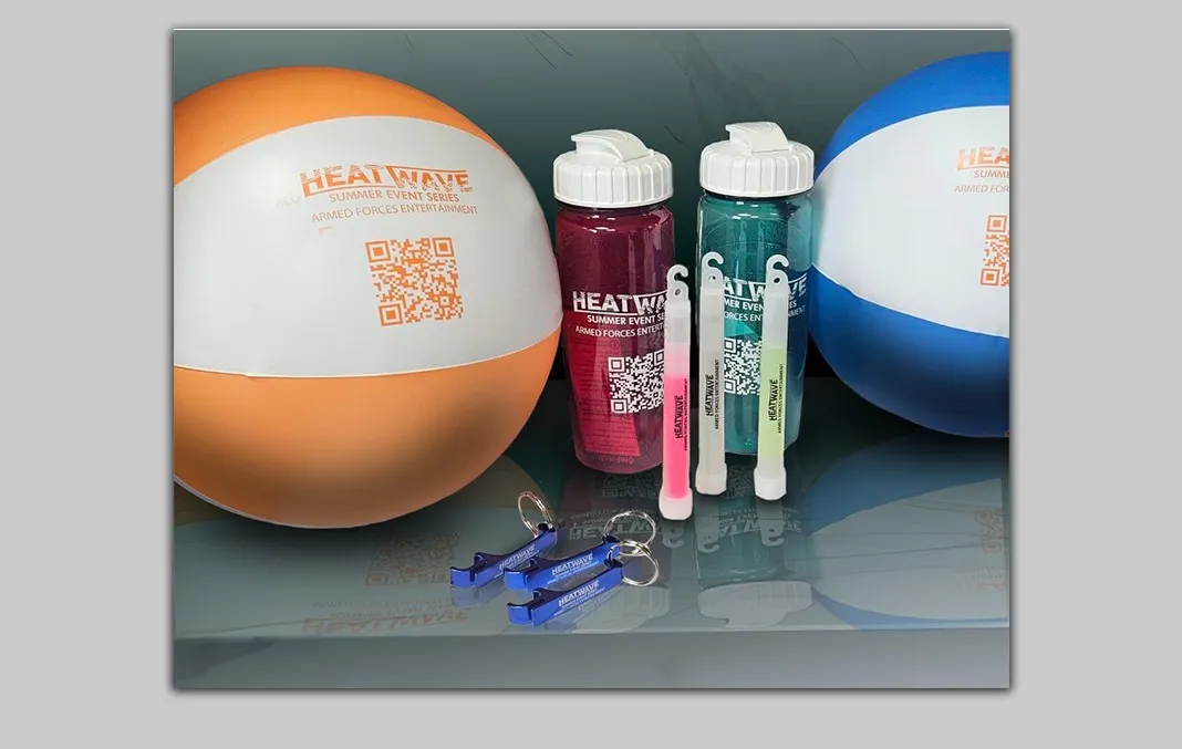 Merchandise for the HeatWave Summer Event Series sponsored by the Armed Forces Entertainment includes two beach balls, two waterbottles, three glow sticks (two yellow and one red), and six bottle opener keychains (three in blue and three in red) assembled on a table.