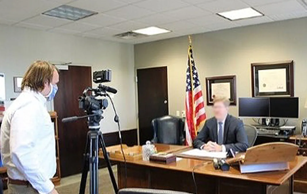 A person in business attire is seated at a desk with an American flag in the background. A cameraman in a mask films from the side.